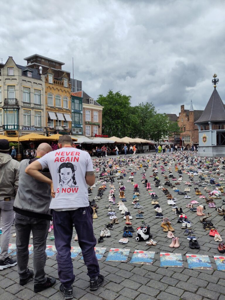 Mensen lezen herdenkingsposters van journalisten die vermoord zijn door Israel. Een persoon heeft een t-shirt aan met een tekening van Hind Rajab, een meisje dat vermoord is door Israel. NEVER AGAIN IS NOW staat er op het shirt.