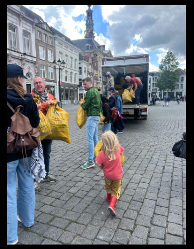 Mensen geven zakken vol met kinderschoenen aan elkaar door door een treintje van mensen te vormen.
Er loopt een jong kind alleen met een grote zak kinderschoenen te sjouwen.