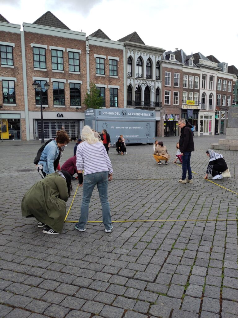 10 a 15 mensen die met meetlinten en krijt vierkanten met stippen maken waarop later kinderschoenen geplaatst kunnen worden.